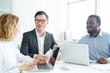 Company of financial analysts or brokers sitting by table and discussing online statistics in office