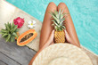 © WHstudio Leushin N - Unrecognizable female tourist rest alone near summer water pool, holds pineapple, surrounded with tropical fruits, enjoys good rest. Suntanned slim woman eats juicy fruit for being healthy and fit
