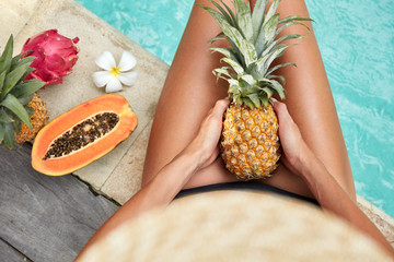 Suntanned female has slender legs, holds exotic pineapple, spends vacation in luxurious hotel, sits near pool with blue water, surrounded with tropical fruits. Summer time and recreation concept