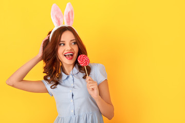 Wall Mural - Young woman studio isolated on yellow wearing bunny ears eating lollipop looking aside