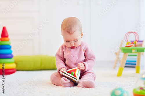 Fototapeta cute little baby girl sitting on the carpet among the development toys, with her first book