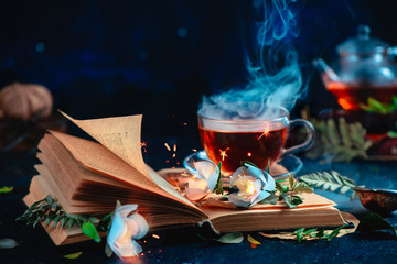 Steaming cup of tea on an open book with magical burning flowers. Fantasy reading concept. Dark still life with copy space.