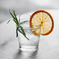 Fresh cocktail with with ice, dried orange slice and fresh tarragon on a white marble table. Macro shot.