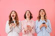 © Drobot Dean - Three lovely young girls 20s wearing colorful striped pyjamas looking upward and using cell phones during happy sleepover, isolated over pink background
