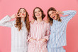 © Drobot Dean - Three cheerful women 20s wearing leisure clothings having fun at slumber party and showing peace signs, isolated over pink background