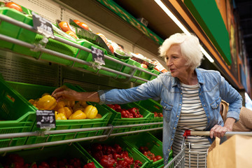 Wall Mural - Portrait of modern senior woman with shopping cart  choosing fruits and vegetables in supermarket while enjoying grocery shopping, copy space