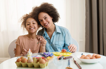 Beautiful African American woman and her daughter coloring Easter eggs at the table  