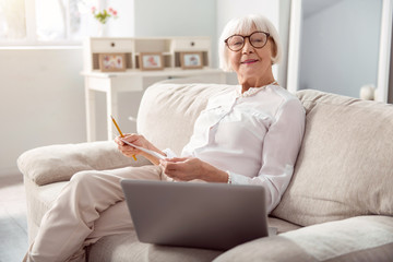 Wall Mural - Brainstorming ideas. Charming elderly woman sitting on the sofa and posing for the camera while developing a project plan and writing down the draft on a sheet of paper