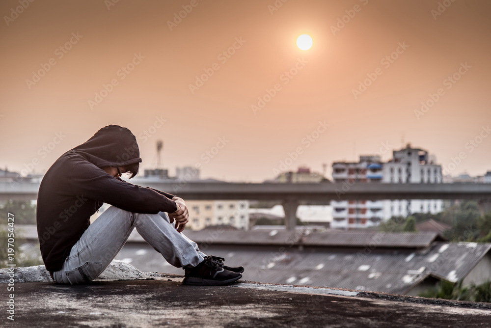 Young Asian man sitting on rooftop of abandoned building with depression stress out during sunset time in the city. Major depressive disorder concept - obrazy, fototapety, plakaty 
