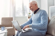 © zinkevych - Mood-boosting chat. Upbeat senior man sitting on the couch and typing an email on the laptop while smiling happily