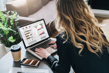 Rear view. Young businesswoman is sitting at table, working on laptop with graphs, charts, diagrams, schedules on screen