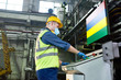 © Seventyfour - Side view portrait of young factory worker wearing hardhat and protective mask operating machine units at modern plant standing by control panel and pushing buttons, copy space