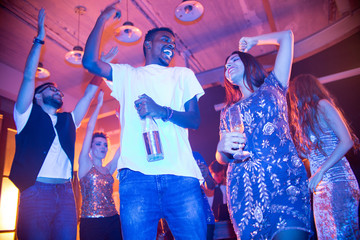 Wall Mural - Low angle view of cheerful friends standing in circle at modern night club and dancing with raised hands, multi-ethnic couple with alcoholic drinks on foreground