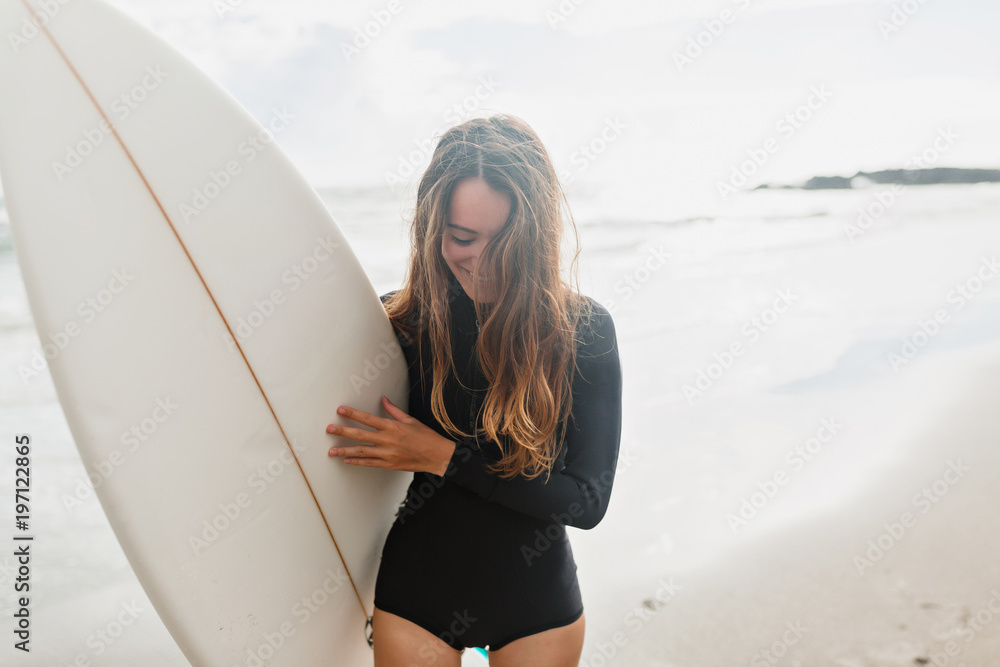 Surfer girl on the beach holding surf board watching ocean waves in sunny day. True happy emotions of young active pretty woman prepare to surfing. Summer vacation lifestyle. - obrazy, fototapety, plakaty 