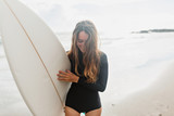 Surfer girl on the beach holding surf board watching ocean waves in sunny day. True happy emotions of young active pretty woman prepare to surfing. Summer vacation lifestyle.