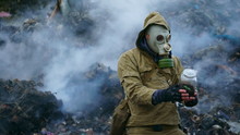 A Man In A Gas Mask Holds A Sprout In The Flask, Against The Background Of Fuming Debris