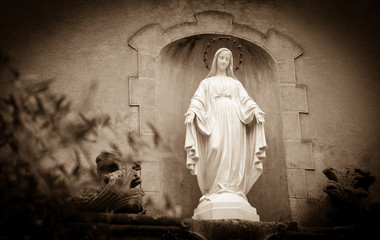 Wall Mural - Statue of Madonna with stars nimbus on front of Cathedral of Notre Dame of Assumption in Entraveaux (France) and olive tree at foreground. Selective focus on statue. Sepia photo.