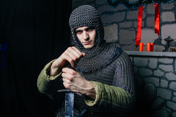 A man with a gun, a stern look. Model man in chain mail hood and chain mail with a sword in his hand. Knight in armor in the castle. Medieval photo illustrations.