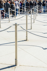 Stainless steel poles linked by grey ropes for queue control at the entrance of a tourist site, with drop shadows on a light ground and blurry people queueing up in the background.