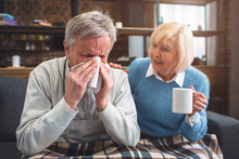 This Ill Man Is Sneezing Into The Napkin. His Wife Is Sitting Besides Him And Advising To Drink Hot Tea From The Cup. She Is Caring About Her Husband.