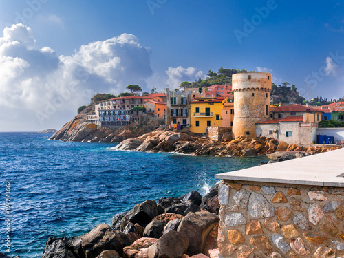 Cuadros en Lienzo Perfect tiny seaside village of Giglio Porto with multi colored houses, an ancient defensive tower and a rocky coastline against a deep blue Mediterranean sea