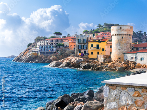 Fotomural The perfect tiny seaside village of Giglio Porto with multi colored houses, an ancient defensive tower and a rocky coastline against a deep blue Mediterranean sea