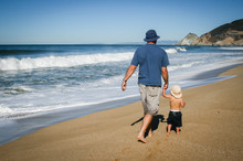 Father And 2 Year Old Son Walking Along The Sand At The Beach In California On A Sunny Day From Behind