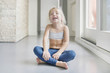 © R - Smiling little blond girl in white top and blue leggings sitting on the floor in a room with crossed legs