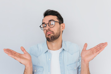 Sticker - Caucasian unsure man make gestures doubtfully with hands with copy space. Uncertain young stylish stubble male with trendy round glasses wears demin blue shirt, shrugs shoulders being puzzled,confused