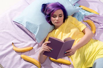 Wall Mural - high angle view of young woman lying in bed with bananas and reading book