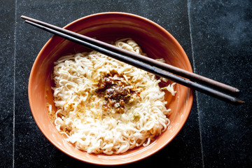 Noodle the sprinkling medium and hot water on orange Bowl. There are chopsticks placed on top. Put on a black tile