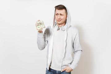 Young handsome male student in t-shirt, blue jeans and light sweatshirt with hood with headphones holds two one-dollar bills and perplexedly looks isolated on white background. Concept of emotions