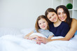 © Maksymiv Iurii - Beautiful mom and daughters are hugging, looking at camera and smiling while sitting on bed at home