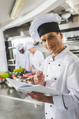 Wall Mural - smiling chef holding recipe book and looking at camera at restaurant kitchen