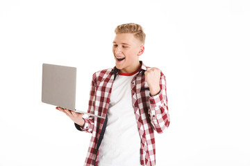 Canvas Print - Portrait of smiling smart teenage boy wearing plaid shirt and braces clenching fist like lucky guy or winner using silver laptop, isolated over white background