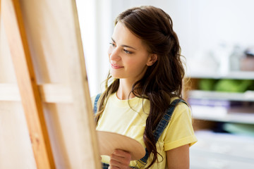 Wall Mural - art school, creativity and people concept - happy smiling student girl or young woman artist with easel and palette painting at studio