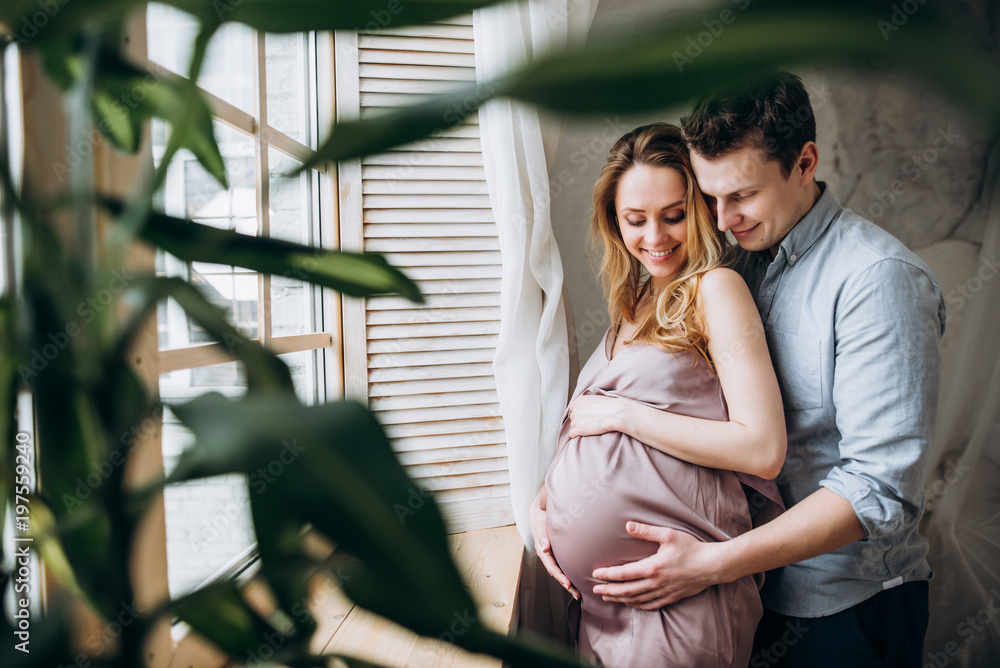 Image of a happy beautiful couple who are waiting for the birth of their baby, they standing by the window and husband hugs their wife with love - obrazy, fototapety, plakaty 