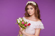 © Надежда Манахова - A beautiful young woman stands on a purple background, wearing a white dress and a wreath of flowers on her head, and a bouquet of tulips. International Women's Day, March 8.