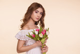 Fototapeta Tulipany - Beautiful young woman stands on a beige background, in a white dress and a bouquet of tulips. International Women's Day, March 8. Copy-space.