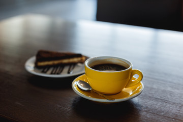 Wall Mural - A horizontal image of a yellow cup of black coffee and a plate with a piece of chocolate cake. Bokeh background.