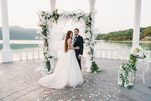 Attractive Groom And Bride At Wedding Day Ceremory With Arch And Lake On Background Stands Together. Beautiful Newlyweds, Young Woman In White Dress And Long Hairs, Men In Black Suit. Happy Family