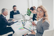 © deagreez - Portrait of smart, concentrated, charming, stylish woman sitting with partners together at desktop in workplace, workstation looking at camera having clipboard with diagrams in hands