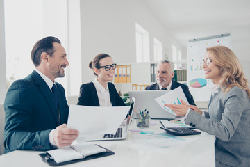 Portrait of four, financial,  stylish, modern business persons in suits gathering in work place station, sitting at desk discussing new project, speaking, having fun, using computers