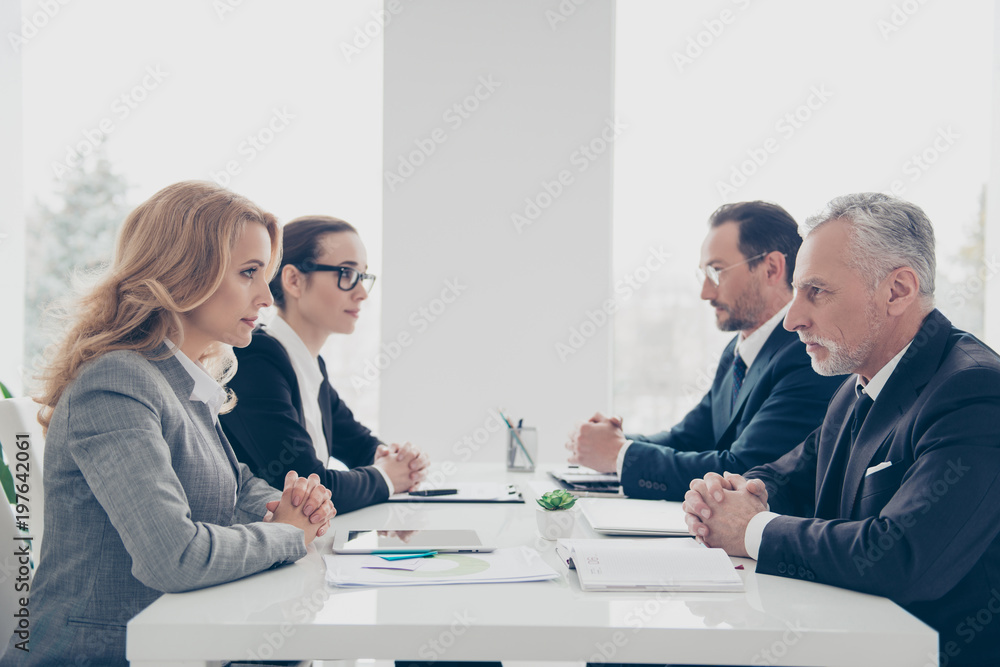 Business competition, four stylish attractive business persons in suits having disagreement and conflict, sitting together at desktop in front of each other, face to face with disrespect expression - obrazy, fototapety, plakaty 