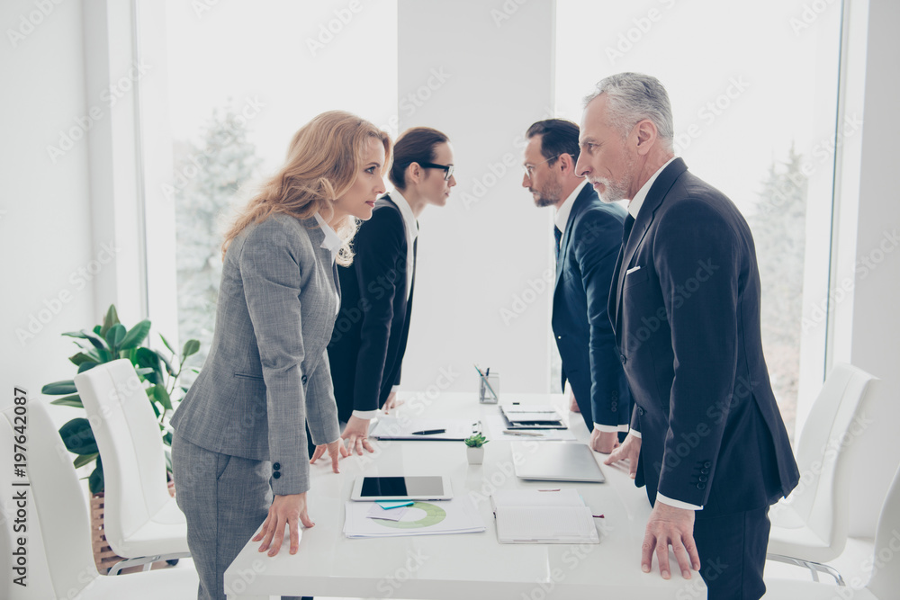 Business competition, four stylish business persons in suits having disagreement and conflict, standing together near desktop in front of each other, face to face with disrespect expression - obrazy, fototapety, plakaty 