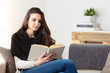 © guerrieroale - Woman reading a book sitting on a couch at home