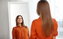 Wardrobe, Fashion, Style And People Concept - Happy Woman In Orange Shirt Looking At Mirror Reflection At Home Or Clothing Store Dressing Room