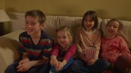 group of children sitting on a sofa and eating popcorn while watching tv. dolly