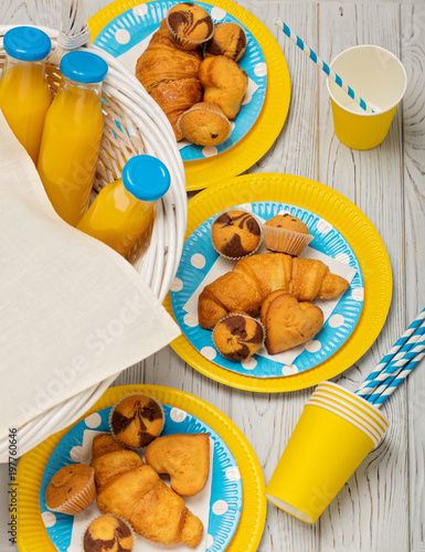 Zdjęcie XXL Letni piknik. Słodki piknik - sok pomarańczowy i babeczki, rogaliki i ciastka na żółtych i niebieskich naczyniach jednorazowych.