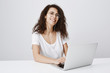 © Cookie Studio - Catching break to talk with coworker during worktime. Happy charming curly-haired female freelancer in glasses looking aside, laughing while working with notebook and listening music in earbuds